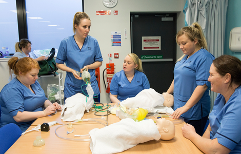 a group of nurses sitting and standing around a table that has the dummy of a newborn baby on it, discussing things and providing it care