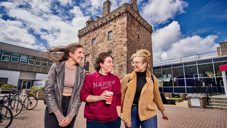 A group of three students walking away from the Merchiston tower under a blue sky.