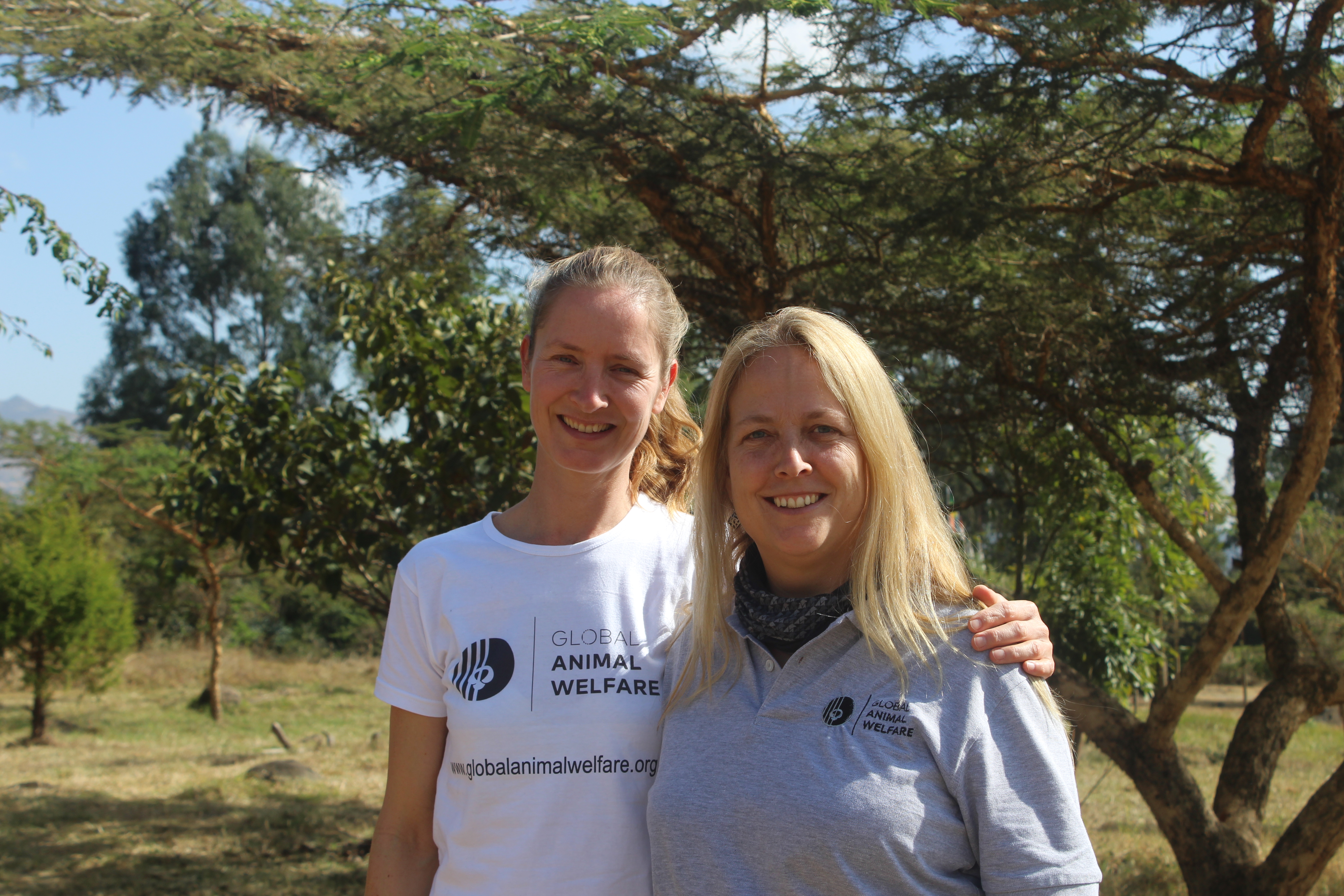 Two women standing in the sunshine, with trees behind them