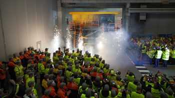 A large group of people wearing hi-vis jackets with volvo on the back, gathered around a vehicle, watching and clapping.