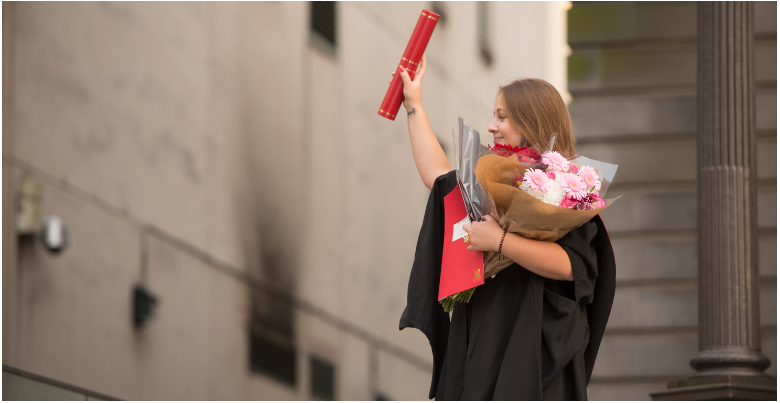 A proud student on graduation day, holding a bouquet of flowers in one hand and a diploma in the other