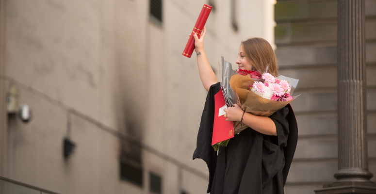 A proud student on graduation day, holding a bouquet of flowers in one hand and a diploma in the other