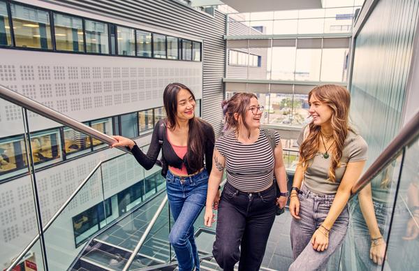 Three students from Edinburgh Napier University conversing while they ascend the main staircase at the Sighthill campus on a sunny day. 