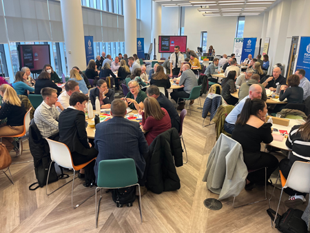 A conference-style event with a room full of people sitting around tables 