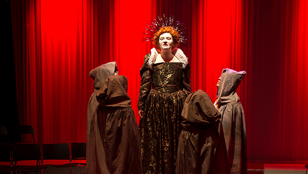 A student actor playing a red haired royal Tudor era character surrounded by hooded figures.